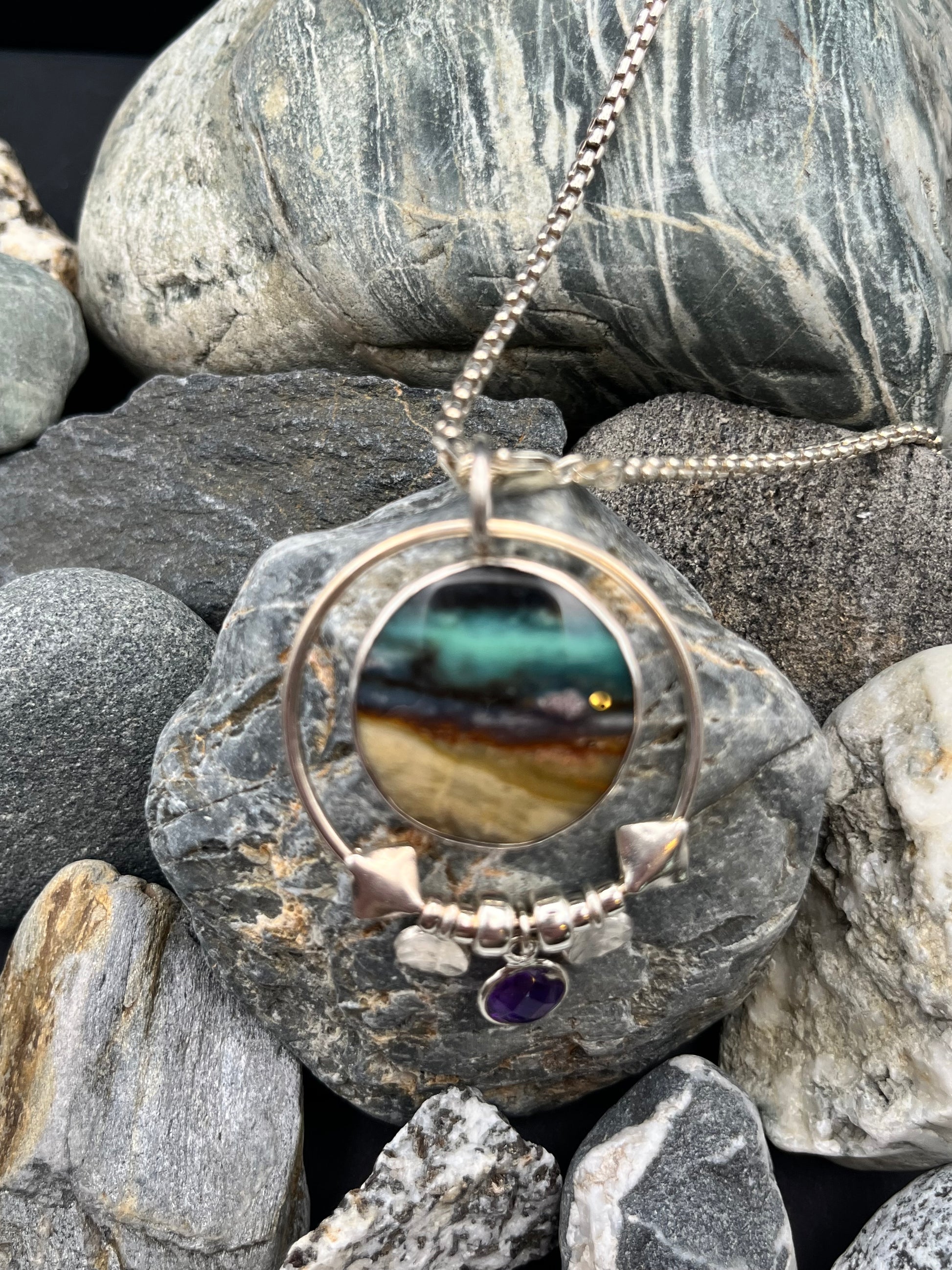 Silver necklace with a round pendant featuring a multicolored stone and purple gemstone, lying on gray stones.