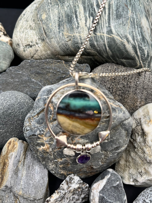 Silver necklace with a round pendant featuring a multicolored stone and purple gemstone, lying on gray stones.
