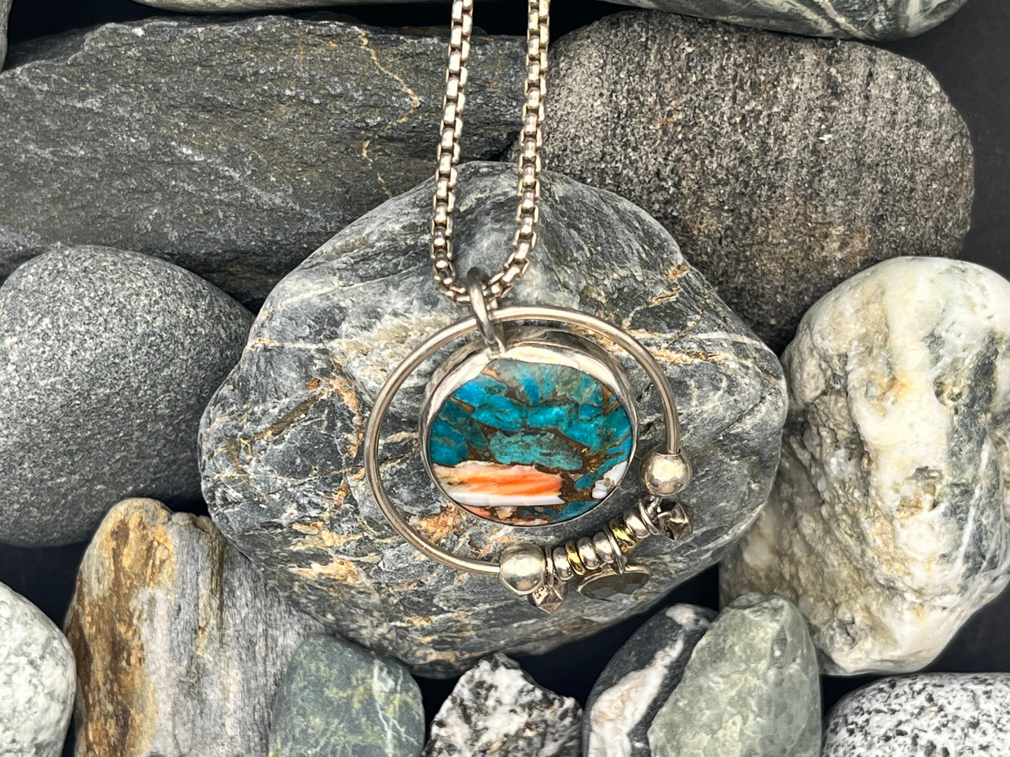Necklace with a circular pendant featuring blue and orange colors, placed on a bed of rocks.